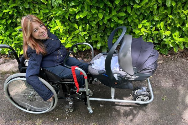 A smiling Natalie using her wheelchair with her baby’s pram attached.